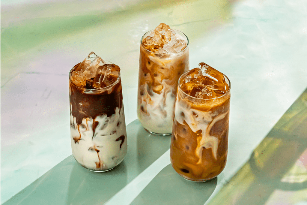 The image shows three tall glasses filled with iced coffee drinks. The drinks have a beautiful swirling effect, with dark coffee blending into creamy white milk, creating an artistic marbled look. The glasses are topped with ice cubes, which sparkle in the light. The surface they are placed on has a soft, pastel-colored background with hints of green, yellow, and pink, adding to the serene and refreshing feel of the image. The sunlight casts gentle shadows, highlighting the textures and layers of the drinks. The overall scene is cool, inviting, and perfect for a hot day.