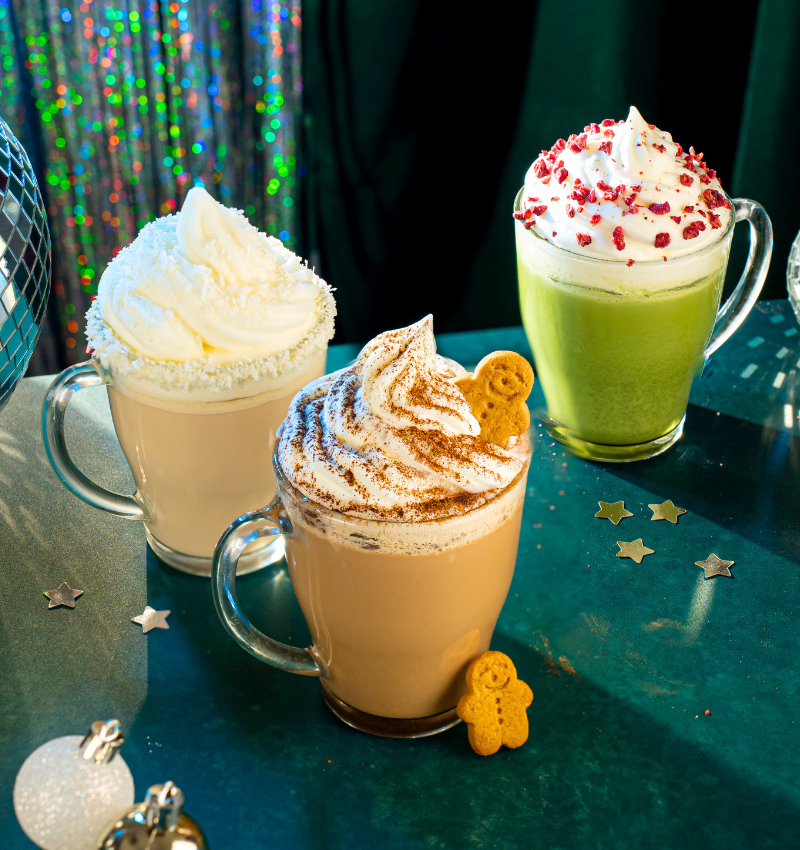 hree festive Christmas drinks in glass mugs on a green surface. From left to right: a white hot chocolate topped with whipped cream, a gingerbread latte topped with whipped cream and cinnamon, and a green matcha latte topped with whipped cream and red sugar crystals. A gingerbread cookie is placed near the gingerbread latte.