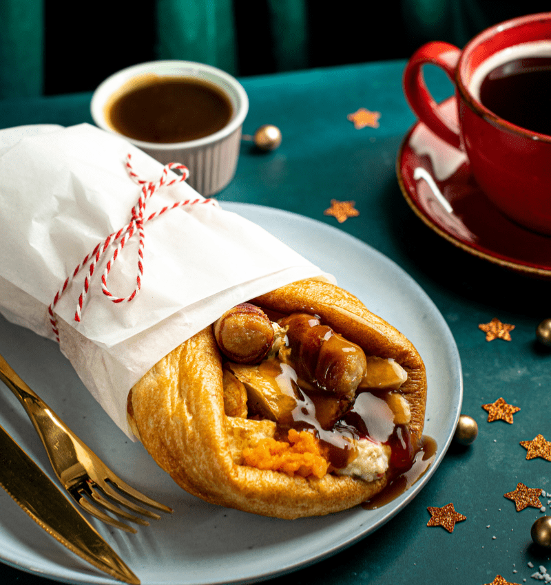 A Christmas dinner wrap, a festive twist on the traditional holiday meal. The wrap is filled with savory stuffing, juicy sausages and a rich, flavorful gravy. The wrap is tied with a red string, adding a touch of holiday cheer. A small bowl of gravy and a steaming cup of coffee complement the wrap, creating a complete and satisfying Christmas meal in a convenient and delicious package.