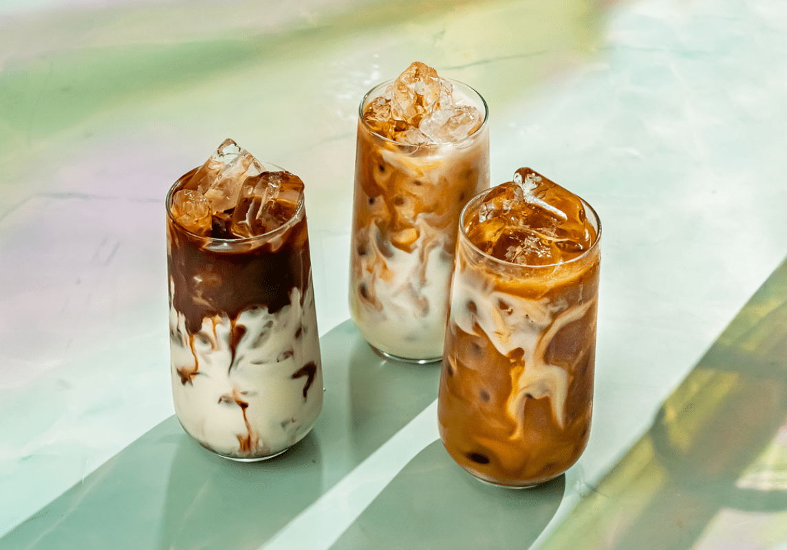 The image shows three tall glasses filled with iced coffee drinks. The drinks have a beautiful swirling effect, with dark coffee blending into creamy white milk, creating an artistic marbled look. The glasses are topped with ice cubes, which sparkle in the light. The surface they are placed on has a soft, pastel-colored background with hints of green, yellow, and pink, adding to the serene and refreshing feel of the image. The sunlight casts gentle shadows, highlighting the textures and layers of the drinks. The overall scene is cool, inviting, and perfect for a hot day.